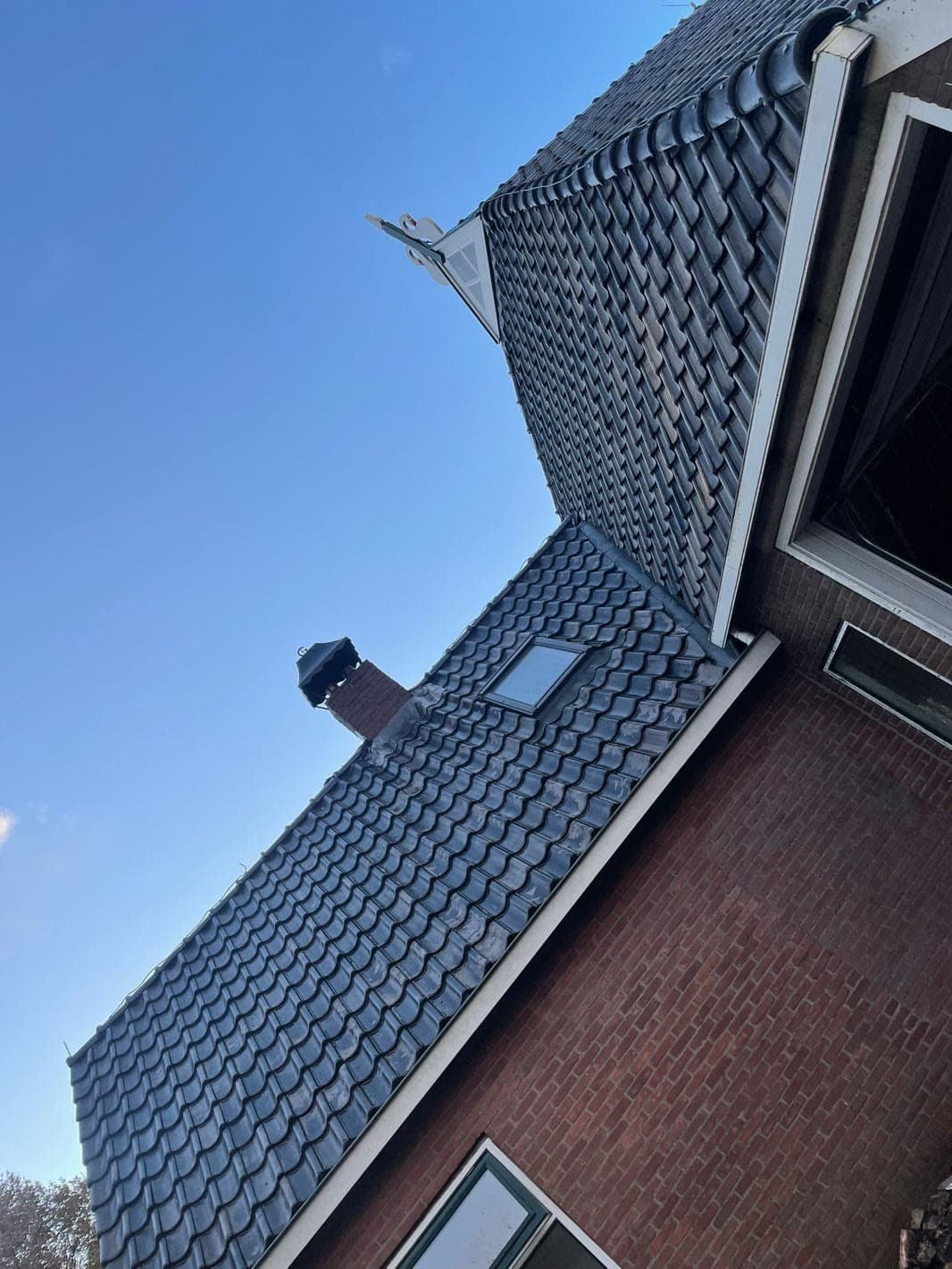 Dakvlak met donkere pannen, nok en dakkapel tegen een blauwe lucht.